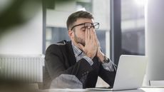 A frustrated office worker puts his face in his hands at his desk.