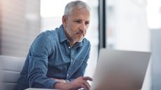 An older man sits at his desk in his office and looks at his laptop. 