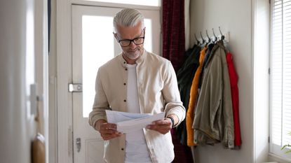 A man with a serious look on his face stands in his foyer and reads a letter he just received.