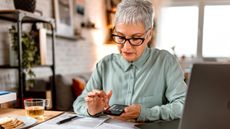 An older woman uses a calculator and looks at paperwork on her kitchen table.