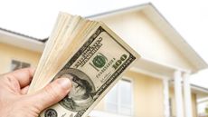 A man holds a stack of cash with a house in the background.